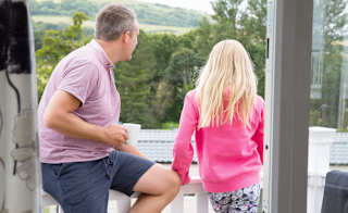 A white man and woman are looking out to the horizon with their back turned to the camer. The man has short grey hair, a blue short sleeved shirt and blue shorts. The woman has long blonde hair, pink long sleeved blouse and patterned trousers. image