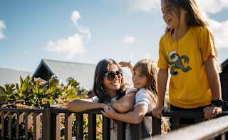 A woman and her two young girls are looking over the wooden balcony. One girl is leaning upward to see more, whilst the other is cuddled around the woman as she smiles at both of the girls. image
