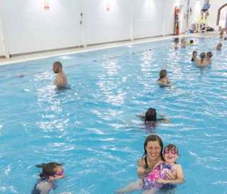 Lots of people of all age ranges swimming in an indoor swimming pool. There is a lifeguard sitting at the site of the pool on a lookout chair.