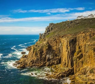 A landscape image of the sea and cliffs located at Lands End. A blue sky with minimal clouds sets the scene with the Land's end tourist building seen in the top right of the image. image