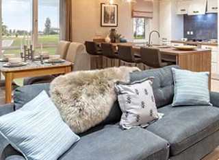 An open-plan view of a holiday caravan. In the foreground is a grey sofa with cushions and a throw. Behind the sofa in the background is a table set for four people and a breakfast bar looking into the kitchen. image