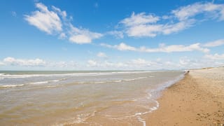 A landscape view of a sandy stoney beach and the sea. The waves are rolling into shore. The sky above is soft blue with multiple clouds.