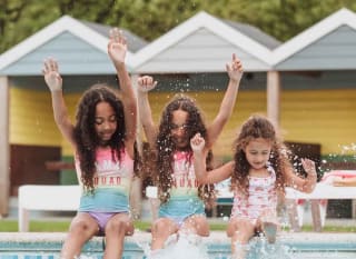 Three girls, each with long brown curly hair and pink swimsuits, are splashing and paddaling on the edge of our outdoor pool at Newquay Bay, Cornwall image