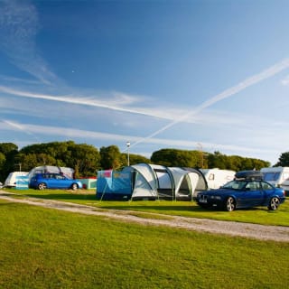 A row of pitched tents, caravans and cars are parked up on a grass parking spot on a sunny day.