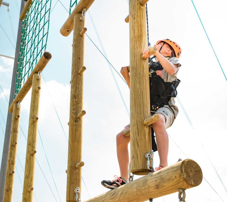 A pre-teen child is climbing on a wodden structure with a bright orange helmet, torso harness and a pully rope system. The child is looking up to judge his next move as he accends higher on the structure. image