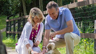 A mature man and woman are petting a light brown and white fluffy dog. Both the man and woman are kneeling down and smiling.
