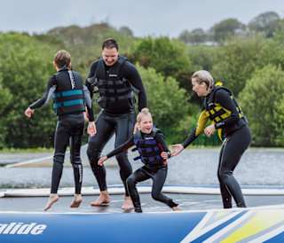 A young family wearing life jackets and full wetsuits balancing on a inflateable in a body of water at one of our holiday parks.