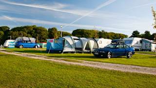 A row of pitched tents, caravans and cars are parked up on a grass parking spot on a sunny day.