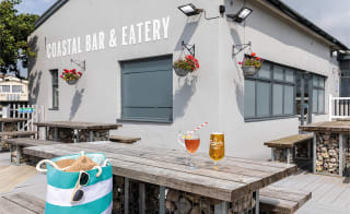 The exterior seating area of The Coastal Bar & Eatery at Bude Holiday Park. There are two drinks on the table and a striped bag on the seat. image