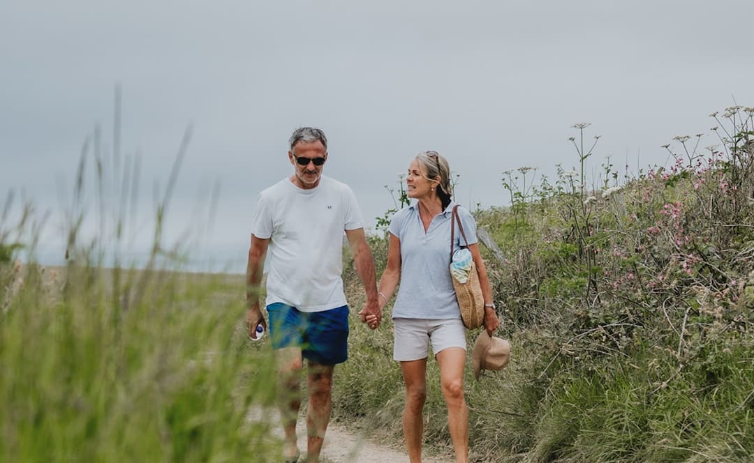 A white, middle-aged couple walking hand in hand down a country path. The man is wearing a white t-shirt with blue shorts. The woman is wearing a light blue polo shirt with cream shorts, whilst holding a weaved hat in her hand. image