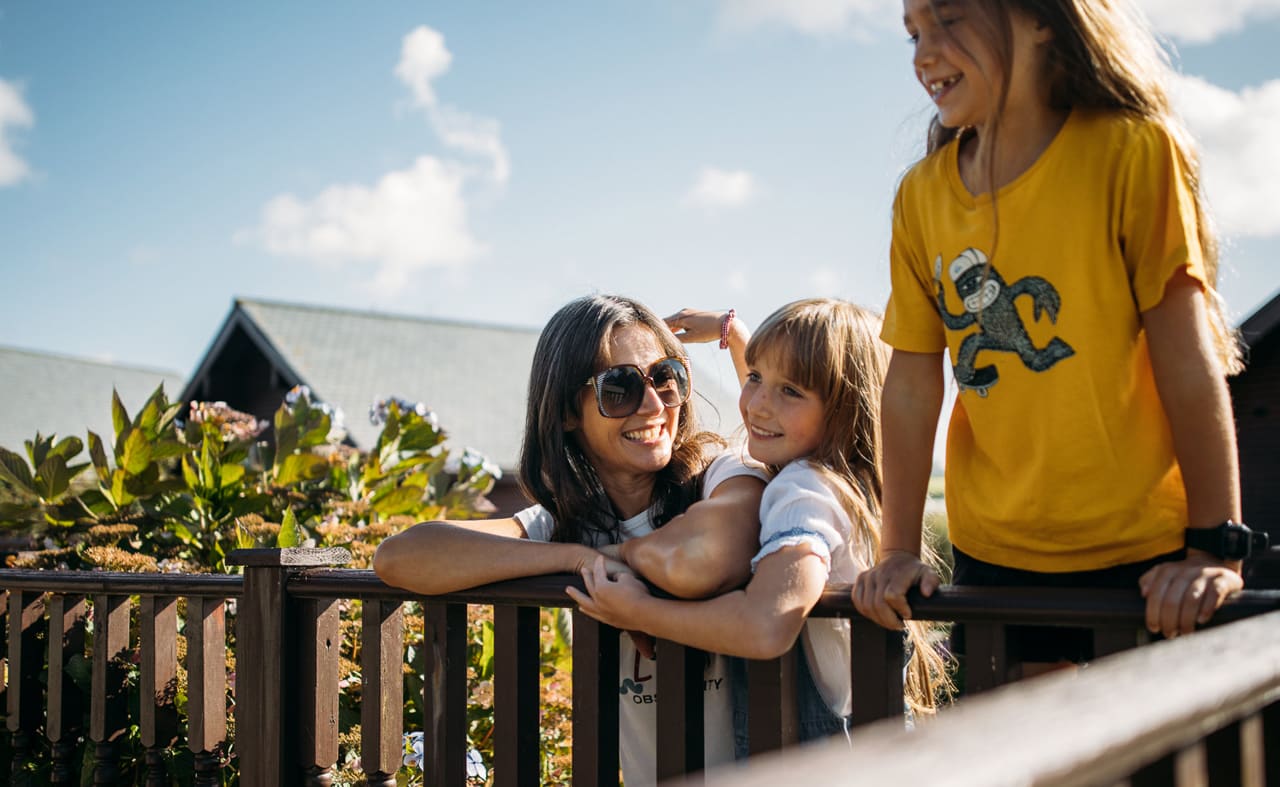 A family enjoying their stay at one of our holiday parks. There is an adult woman and two young girls. One of the girls is hugging the woman, the other is leaning over a banister. All three have wide smiles. image