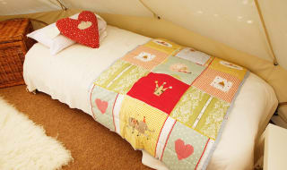 A photo of the bedroom area within a off-white canvas bell tent. The bed is a single camping bed with a cream duvet, pillow and red spotted heart-shaped cushion. image