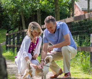 A mature man and woman are petting a light brown and white fluffy dog. Both the man and woman are kneeling down and smiling.