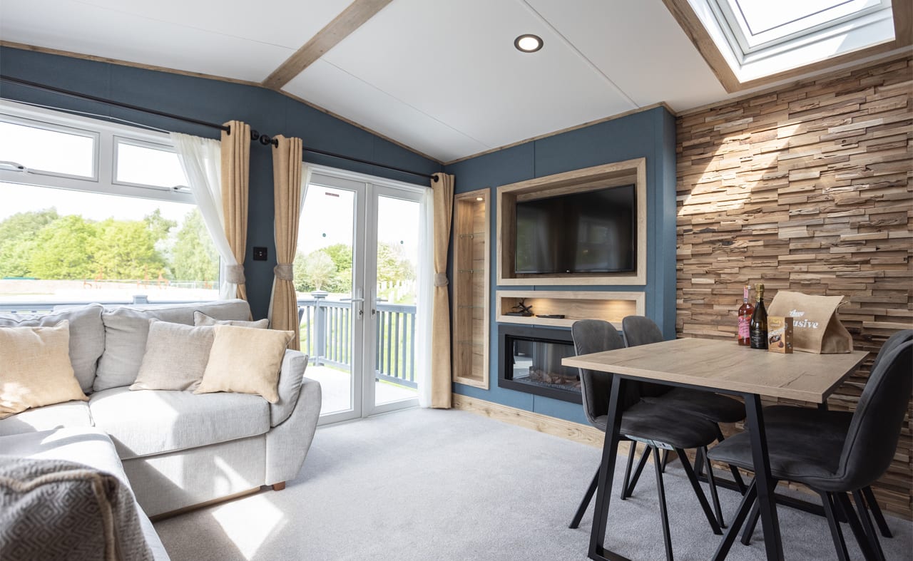 The inside of one of our static accessible caravans. There is a grey sofa and a dining area, along with a TV and fireplace. The large double doors are letting in sunlight. image