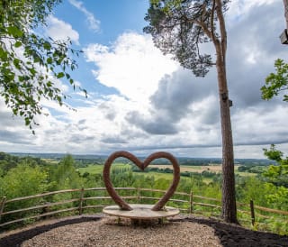 A panoramic view of the countryside, with a viewing area in the foreground. The bench features a wooden heart arch.