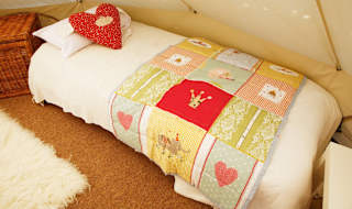 A photo of the bedroom area within a off-white canvas bell tent. The bed is a single camping bed with a cream duvet, pillow and red spotted heart-shaped cushion. image