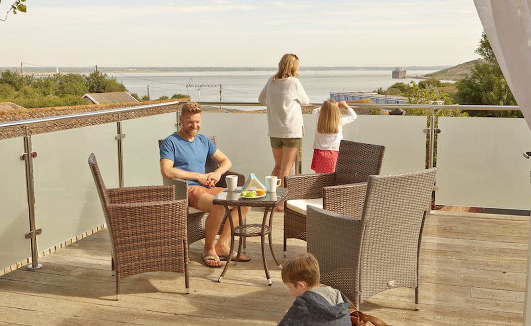 A family of four on the balcony of their holiday home in The Bay Colwell. There are two adults, a young boy, and a young girl with strawberry blonde hair, and all dressed in summer attire. image