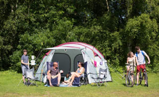 A pitched up family tent with a man, woman and baby sitting in the porch area of the tent. A teen boy and girl are cycling by on a hot sunny day. image
