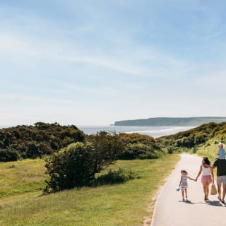 A young family are walking along a dry dirt path which is situated between rolling green hills on a hot summer's day. image