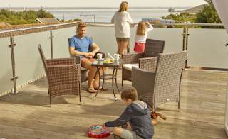 A family of four on the balcony of their holiday home in The Bay Colwell. There are two adults, a young boy, and a young girl with strawberry blonde hair, and all dressed in summer attire. image