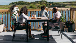 A family sat outside on the decking of their holiday home on a sunny day at The Bay Filey, Yorkshire, overlooking the lake and woodland views.