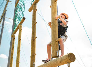 A pre-teen child is climbing on a wodden structure with a bright orange helmet, torso harness and a pully rope system. The child is looking up to judge his next move as he accends higher on the structure. image