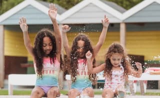Three girls, each with long brown curly hair and pink swimsuits, are splashing and paddaling on the edge of our outdoor pool at Newquay Bay, Cornwall image
