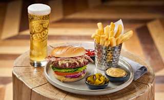 On the place is a stacked bacon cheeseburger with lettace, tomato and sliced red onion. Next to the burger is cooked chips and ketchup, pickles and burger sauce. Next to the plate on the left is a tall glass of larger with a thick, creamy head. image