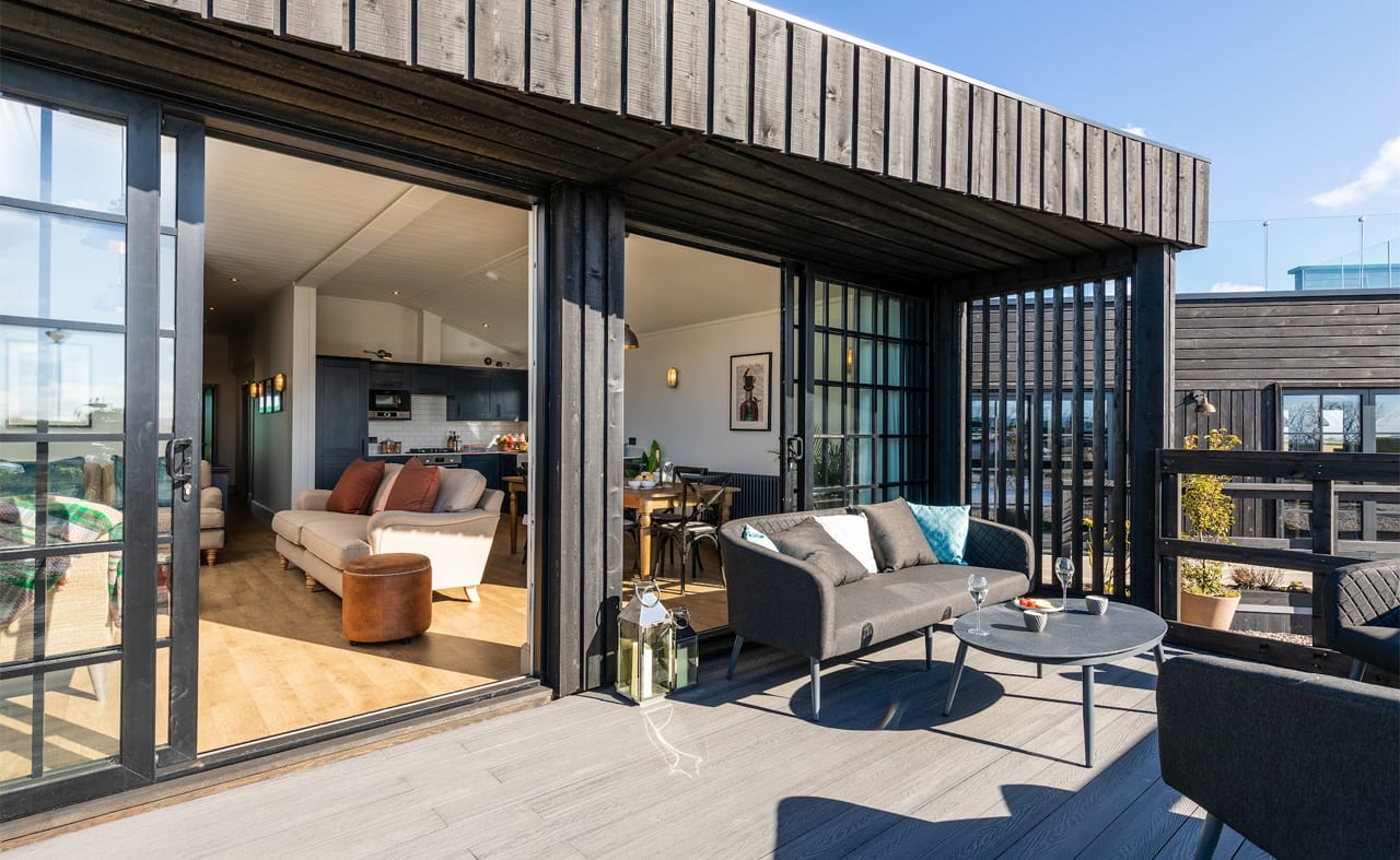 An open-plan barn, looking from the outside in. Starts with the outside balcony looking into the lodge where a cream sofa can be seen with a hint of the kitchen in the distance. image