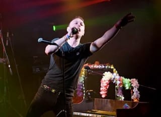 An adult male on a stage singing at a rock gig. He's got short light brown hair, a black tee and dark jeans singing into the microphone and pointing out to the crowd. image