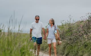 A white, middle-aged couple walking hand in hand down a country path. The man is wearing a white t-shirt with blue shorts. The woman is wearing a light blue polo shirt with cream shorts, whilst holding a weaved hat in her hand. image