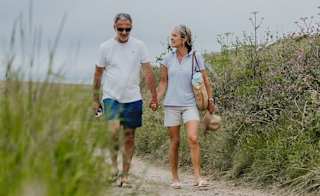 A white, middle-aged couple walking hand in hand down a country path. The man is wearing a white t-shirt with blue shorts. The woman is wearing a light blue polo shirt with cream shorts, whilst holding a weaved hat in her hand. image