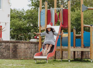 A wooden children's adventure playground outside on some trimmed grass at Newquay Bay, Cornwall. A girl wearing a white hat and a checkered dress is sliding down a red slide. image