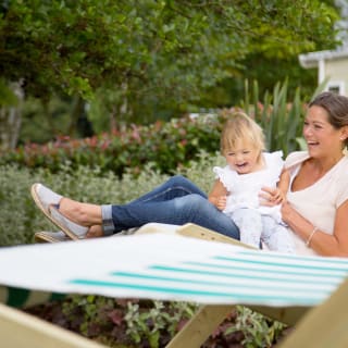 An adult white woman and young child are outside sitting on a wooden deck chair. The child is sat on the lap of the woman and has a gleeful expression. The woman is suporting the child upright and is smiling.
