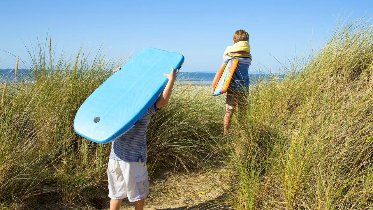 Two children walking across the sand to the sea holiday two body boards on a sunny day.