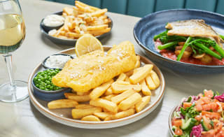 A close up of fish and chips on a wooden table with various other dishes around it. image