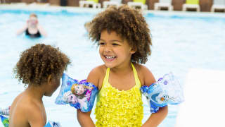 Two young children paddling in an outdoor pool. One is wearing a bright yellow swimming costume with armbands and the other is mostly out of shot but some of their curly hair is in frame.