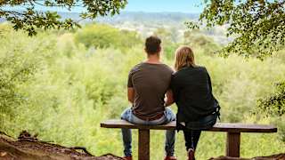 A couple sat on a bench looking out to stunning woodland views on a summers day.