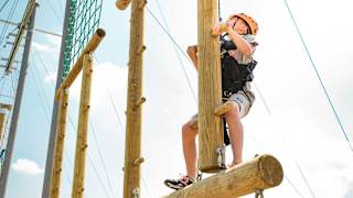 A pre-teen child is climbing on a wodden structure with a bright orange helmet, torso harness and a pully rope system. The child is looking up to judge his next move as he accends higher on the structure.