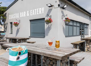 The exterior seating area of The Coastal Bar & Eatery at Bude Holiday Park. There are two drinks on the table and a striped bag on the seat. image