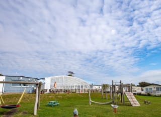 The outdoor adventure playground at our Essex holiday park, Mersea Island. There is views of the indoor swimming pool conservatory and some caravans in the background. image