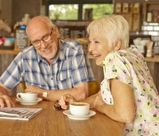 An elderly couple enjoying a coffee at The Bay Colwell. The couple are laughing and smiling, and both have short grey hair. They each have a full mug of coffee, and in the background there is a barista making coffee and customer walking.