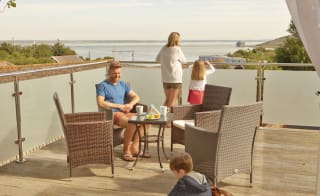 A family of four on the balcony of their holiday home in The Bay Colwell. There are two adults, a young boy, and a young girl with strawberry blonde hair, and all dressed in summer attire. image