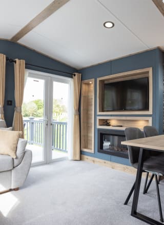 The inside of one of our static accessible caravans. There is a grey sofa and a dining area, along with a TV and fireplace. The large double doors are letting in sunlight.