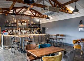 A restaurant bar area, with grey walls and wood panelling. Four tables with chairs can be seen in the image. The bar is stocked with bottles on the back wall with a mirror hanging above. image