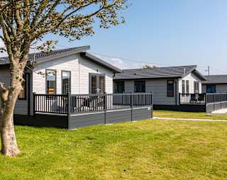Our amazing lodges at one of our holiday parks. There are three luxury lodges surrounded by green grass, and the sun is shining down. image