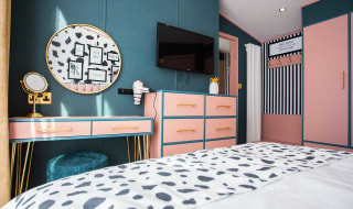 In the double bedroom is a large light pink and gold vanity table with a large circular mirror on the wall and a flat screen TV hanging on the wall next to it. image