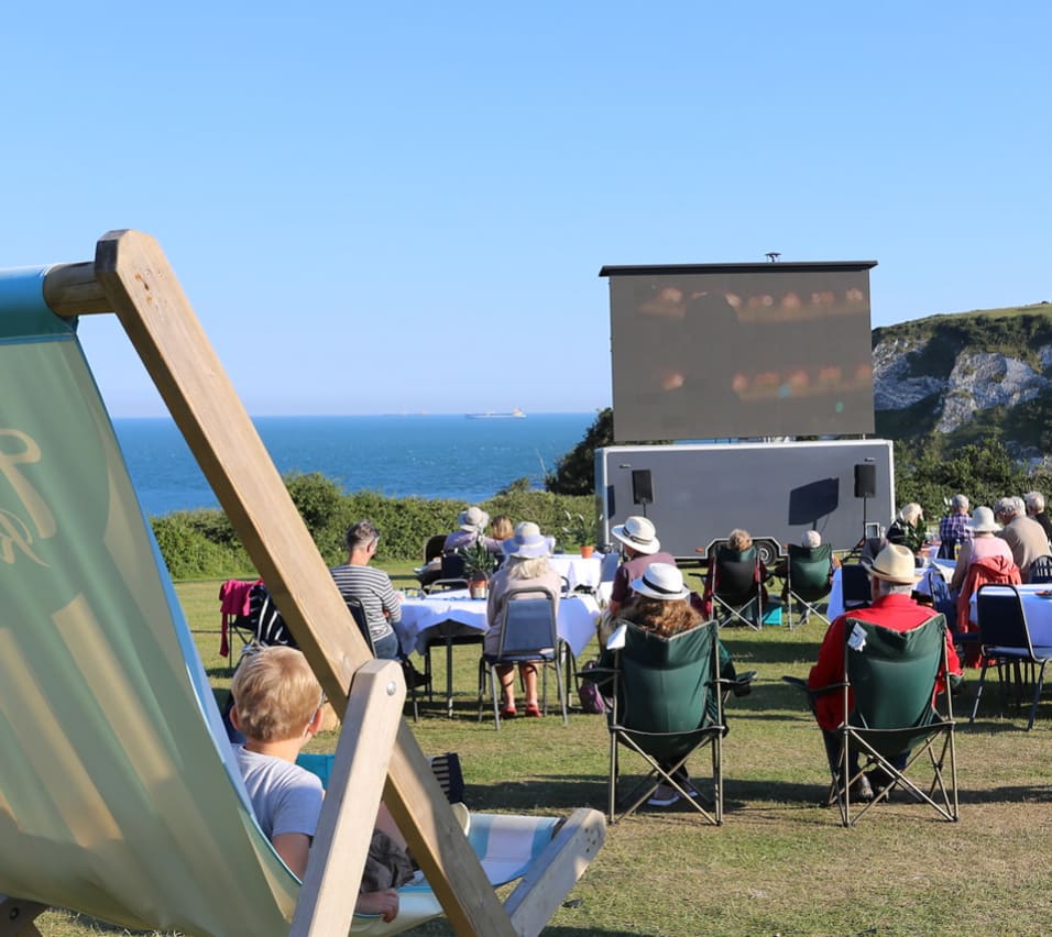 A group of people enjoying the outdoor cinema at our stunning holiday park on the Isle of Wight, Whitecliff Bay. There are views of the sea in the background on a summers day. image