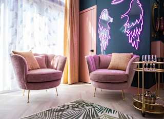 The stunning lounge area in The Esme Compact. There are two plush velvet chairs, a gold drinks trolley and some pink neon lighting in the shape of angel wings. image