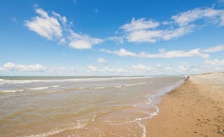 A landscape view of a sandy stoney beach and the sea. The waves are rolling into shore. The sky above is soft blue with multiple clouds. image
